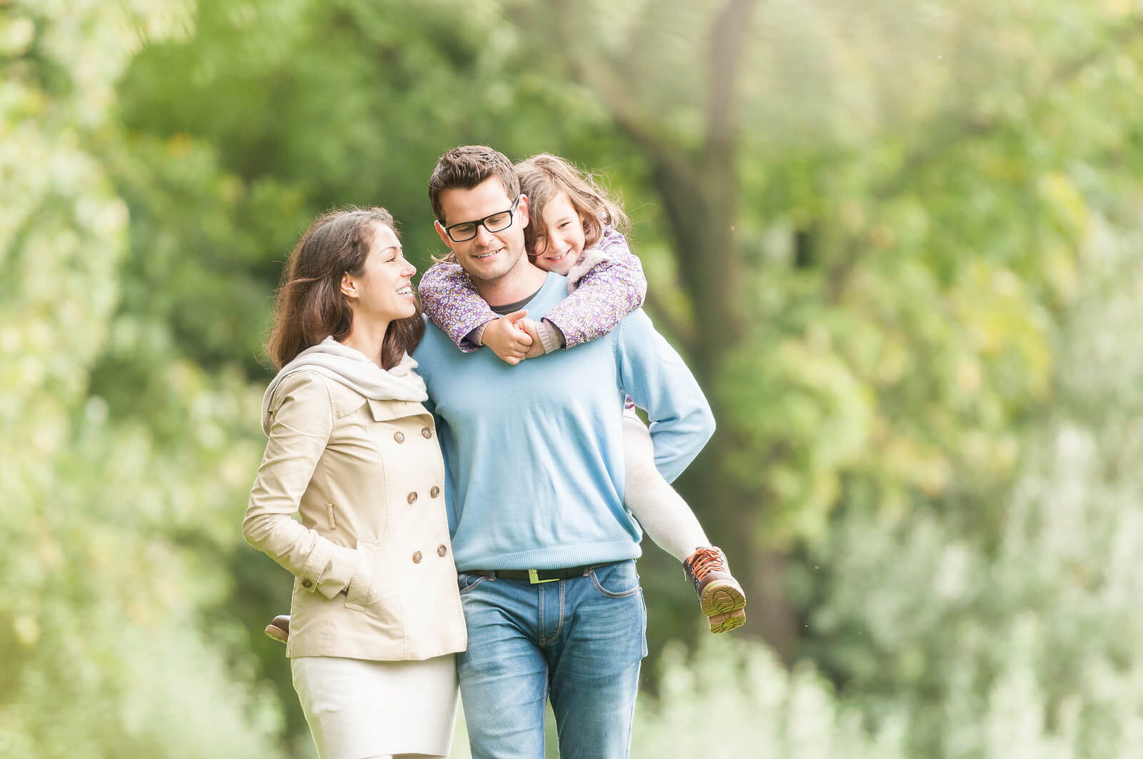 Young happy family of three having fun together outdoor. Pretty little daughter on her father back. Parents and girl look happy and smile. Happiness and harmony in family life. Family fun outside.